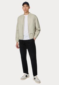 Lichtbeige bomberjack met geribbelde manchetten, witte t-shirt, zwarte broek en grijze sneakers met zwarte accenten, gedragen door een staande model.