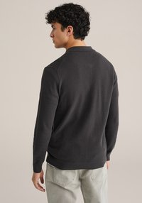 Temno sivi pleteni pulover z rebrastim ovratnikom in robom. Ima teksturirano tkanino in prilagojen kroj, nošen z lahkotnimi svetlimi hlačami.