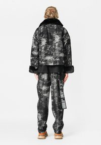 Abrigo de piel con un patrón en negro y gris, con un cuello ancho, combinado con pantalones a juego que tienen una etiqueta lateral y un corte holgado. Los zapatos son de color marrón.