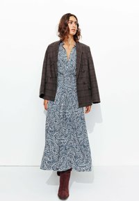 Robe maxi à motif paisley bleu avec un décolleté en V, taille cintrée, associée à un blazer à carreaux marron foncé et des bottes en suède marron jusqu'aux genoux.