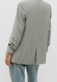 Personne portant un blazer gris clair sur une chemise en jean avec un jean large bleu délavé, debout devant un fond clair uni.