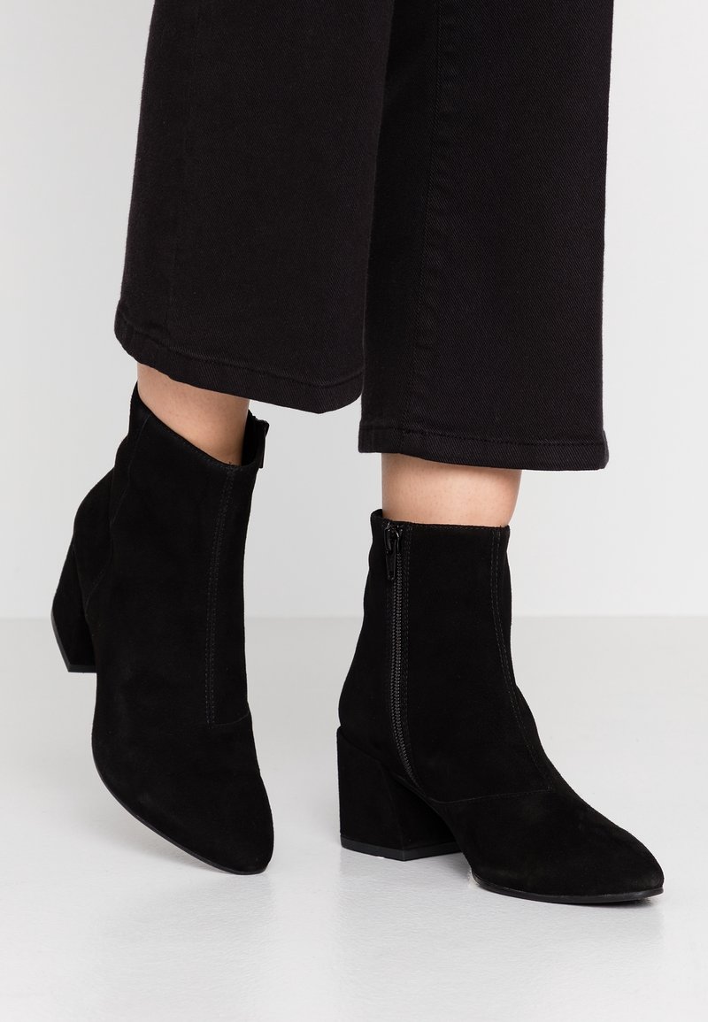 Pieds portant des bottines en daim noir à talons blocs et fermetures éclair latérales, associées à un pantalon noir raccourci sur un fond clair.