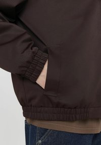 Veste isolante marron avec des poignets et un ourlet élastiques, dotée d'une poche latérale et d'un tissu lisse et texturé. Portée sur un haut marron et un jean bleu.