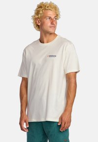 Camiseta de algodón de manga corta en color crema con un corte relajado, que presenta un pequeño logo azul y rojo en el lado izquierdo del pecho.