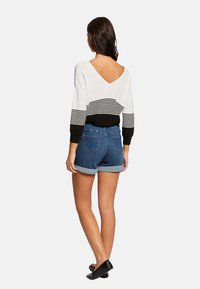 Maglione a righe bianche e nere con scollatura a V sul retro, abbinato a pantaloni shorts in denim blu con orlo arrotolato e tasche posteriori, indossato con ballerine nere.