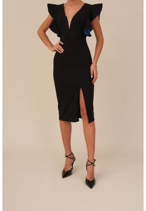 Woman wearing a black knee-length dress with deep V-neck, ruffled cap sleeves, front slit, and black strappy high heels.