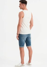 Witte mouwloze tanktop en blauwe denim shorts met omgerolde zoom. Schoeisel is witte sneakers. Achtergrond is eenvoudig wit.