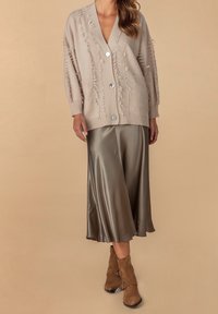 Beige Cardigan mit fransiger Textur, großen Knöpfen und Taschen, kombiniert mit einem fließenden, glänzenden olivgrünen Satinrock und Wildleder-Ankle-Boots.
