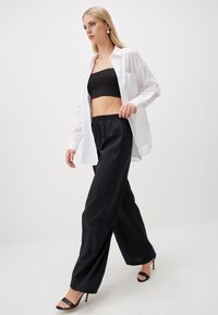 Zwarte crop top gecombineerd met loszittende zwarte broek; model draagt een witte button-up shirt en zwarte hoge hakken. Gladde stof, relaxte fit.