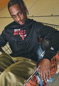 Haut à capuche noir avec le logo "Chicago Bulls" en rouge, associé à un pantalon vert olive, assis dans un chariot de course en fil métallique avec des ballons de basket.