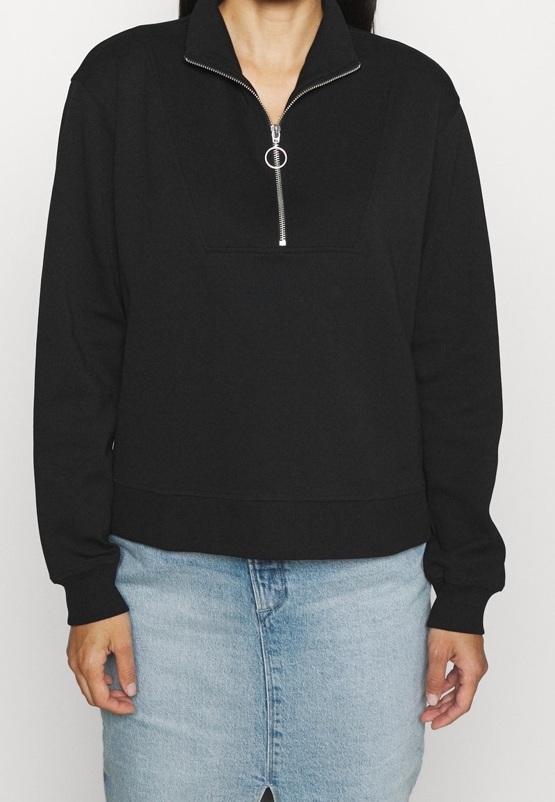 Femme portant un pull noir à manches longues avec une demi-fermeture éclair argentée et un jean taille haute bleu clair.