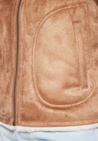 Brązowa kurtka z sztucznego zamszu z zamkiem błyskawicznym, z miękkim białym wykończeniem i półokrągłymi kieszeniami naszytymi z przodu. Gładka faktura na całej powierzchni.