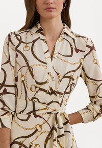 Wrap dress in cream fabric featuring a chain and harness print in brown and gold. Long sleeves with cuffs, tied waist detail.