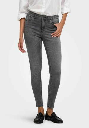 Personne portant un jean gris slim-fit, une chemise blanche retroussée et des mocassins noirs, debout avec une main dans la poche devant un fond uni.