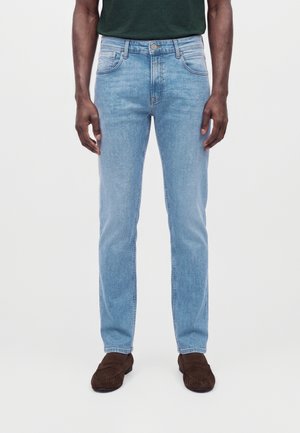 Homme portant un jean slim bleu clair, une chemise vert foncé et des mocassins en daim marron, debout devant un fond uni.