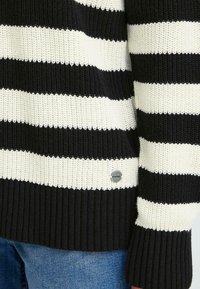 Svart- og hvitstripete strikkegenser med ribbestruktur og en sølvfarget logo. Stoffet har brede horisontale striper og en løs passform.