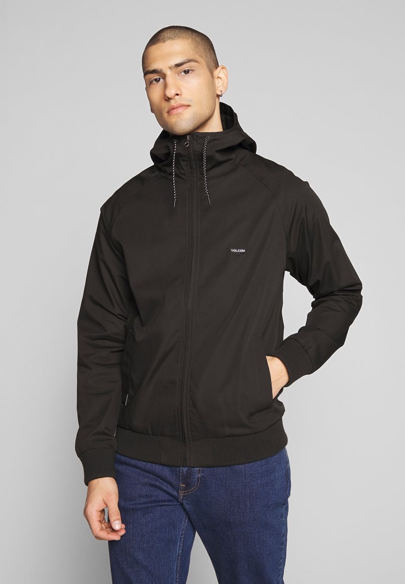 Sort zip-up jakke med hætte, der har en glat tekstur, ribbede manchetter og et lille logo på brystet. Sammen med blå jeans.