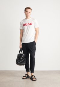 Bílý tričko s červeným logem "HUGO", černé cargo kalhoty, černá taška přes rameno a černé sandály se strapcemi. Hladká tkanina a casual design.