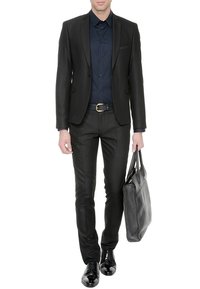 Homme portant un costume noir et une chemise bleu marine, tenant une grande mallette en cuir noir dans sa main droite, debout devant un fond blanc.
