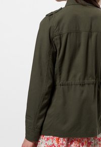 Giacca verde oliva con vita aderente, maniche lunghe e mostrine sulle spalle; tessuto liscio senza chiusure o motivi visibili.