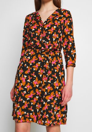 Kvinde iført knælang sort kjole med orange og pink blomsterprint, trekvartlange ærmer og bælte i taljen.