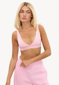 Femme aux longs cheveux blonds portant un haut brassière rose clair et un pantalon de jogging rose assorti sur un fond uni.