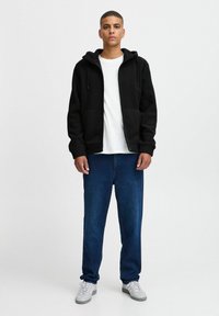 Black zip-up hoodie with a hood, front pockets, worn over a white t-shirt. Paired with loose blue jeans and gray sneakers.