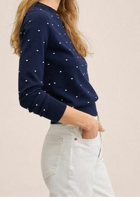 Pull bleu marine à pois blancs, col rond et poignets côtelés, associé à un pantalon de couleur claire. Texture lisse, coupe décontractée.