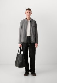 Chaqueta gris texturizada con un bolsillo frontal, camisa blanca, pantalones de mezclilla negros y un bolso tote de cuero negro, de pie sobre un fondo neutro.
