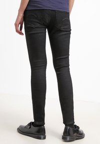 Vaqueros negros ajustados con una textura suave, que incluyen bolsillos traseros y detalles de costura sutiles, combinados con zapatos negros.