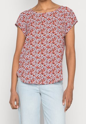 Femme portant un chemisier à manches courtes, ample, avec un motif floral rouge et rose, associé à un jean bleu clair.