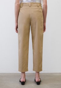 Osoba v béžových zkrácených kalhotách a černých sandálech na vysokém podpatku stojí na světlém šedém podlaze proti bílé zdi.