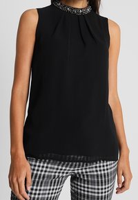 Zwarte mouwloze blouse met een geplooide voorkant en een bewerkte halslijn, met een iets doorzichtige zoom. Gecombineerd met zwart-witte geruite broeken.