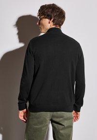 Černý pletený svetr s dlouhými rukávy a vysokým límcem. Má žebrované manžety a spodní lem, nošený přes olivově zelené kalhoty.