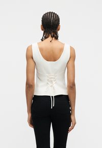 Άτ ομο με πλεξούδες, ντυμένο με λευκό αμάνικο τοπ με δέσιμο δαντέλας στην πλάτη και μαύρο παντελόνι, γυρισμένο προς τα πίσω, μπροστά σε απλό φόντο.