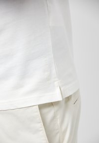 T-shirt en coton blanc avec des fentes sur les côtés, présentant une finition texturée subtile et un logo embossé près de l'ourlet. Associé à un pantalon beige.
