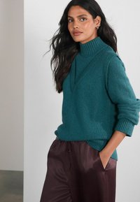 Pulover tricotat în nuanță de verde-albastru, cu guler înalt, având un decolteu în formă de V și maneci cu revere, purtat cu pantaloni de satin maroo închis.