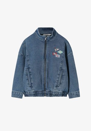 Kind met krullend haar staat glimlachend, draagt een lichtgele trui, spijkerjasje, opgerolde jeans en beige sneakers tegen een beige achtergrond.