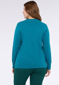 Kobieta z brązowymi włosami, ubrana w turkusową bluzkę z długim rękawem i ciemnozielone spodnie, stojąca plecami do kamery na białym tle.