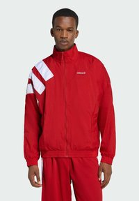 Červená vetrovka s bielymi akcentmi na ľavom pleci a vyšitým logom na hrudi; priliehavé manžety a spodný lem.
