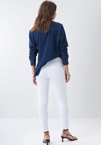 Camisa de mezclilla en azul oscuro con mangas largas y dobladillo trasero curvado, combinada con jeans ajustados blancos y zapatos de tacón negros con tiras.