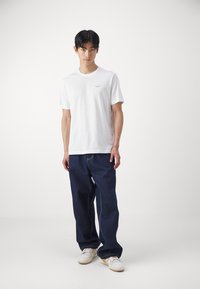 Witte katoenen t-shirt met een klein bedrukt logo, gecombineerd met navy blauwe losvallende jeans en witte sneakers. Minimalistisch ontwerp, ontspannen stijl.