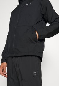 Personne portant une veste Nike noire avec une fermeture éclair et la main dans la poche, associée à un pantalon Nike noir avec une petite étiquette sur la cuisse.