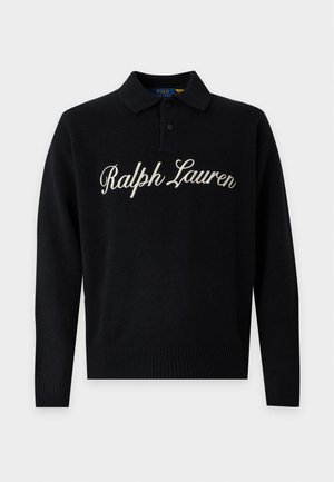 Giovane uomo che indossa una camicia nera Ralph Lauren a maniche lunghe, jeans azzurri a gamba larga e sneaker bianche, in piedi davanti a uno sfondo semplice.