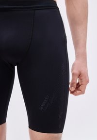 Κοντινό πλάνο ενός ατόμου που φοράει μαύρα μαγιό Speedo jammers με υφασμένο λογότυπο στον μηρό, όρθιο εμπρός σε απλό φωτεινό φόντο.