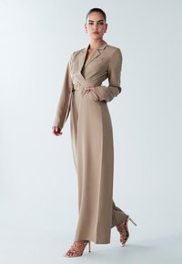 Bēša garš kombinezons ar žaketa stila elementi, jostu pie vidukļa, garām piedurknēm un sānu kabatām. Kombinēts ar zelta krāsas sandālēm ar augstiem papēžiem un siksniņām.