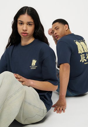 Una donna e un uomo sono seduti per terra indossando magliette blu navy con disegni gialli di gatti della fortuna; la donna è rivolta in avanti, l'uomo di lato.