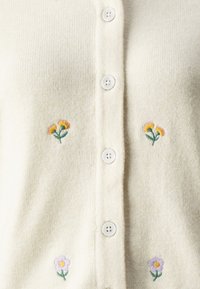 Helles beige Strickjacke mit fünf weißen Knöpfen. Enthält bestickte Blumen: zwei gelbe und rosa Blumenstöcke sowie eine lila Blüte. Weicher Stoff.