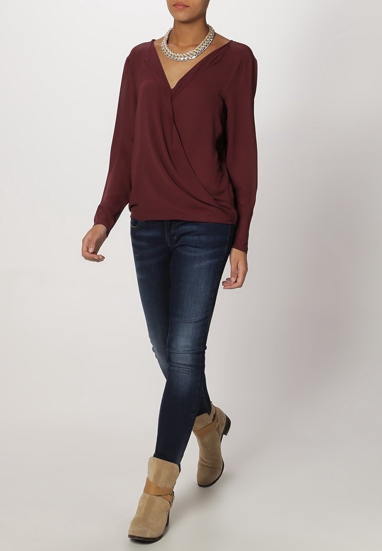 Blusa envolvente de color burdeos con mangas largas y escote en forma de V, combinada con jeans ajustados de un azul oscuro y botas de tobillo beige con detalle de tira. Collar de plata.