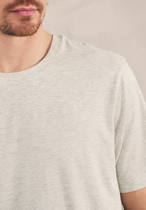 Mand med let skæg iført en lysegrå, fint striped kortærmet T-shirt med et subtilt broderet logo på brystet.